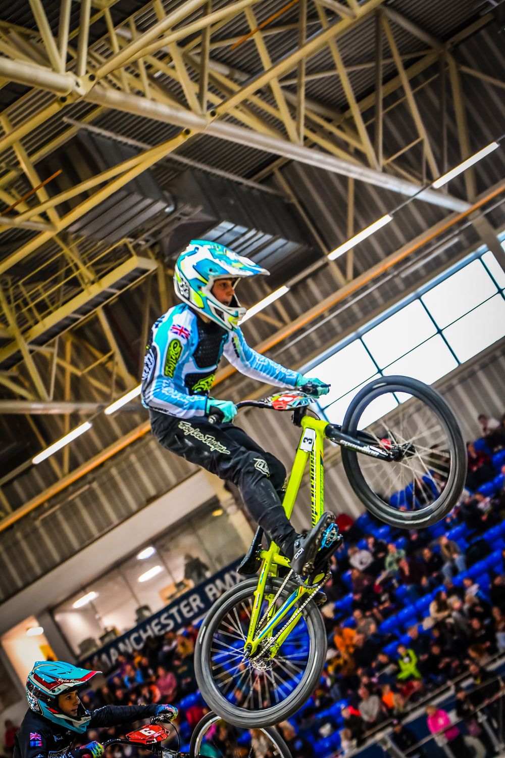 Charlie airborne over a jump on an indoor BMX track, crowd in the stands behind